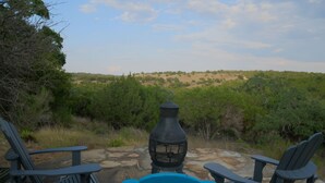 Eine Terrasse mit zwei blauen Adirondack-Stühlen, einem schwarzen Metalltisch und einem schwarzen Laternenlicht. Im Hintergrund eine natürliche Landschaft mit Bäumen und Hügeln.