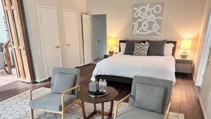 A hotel room with a bed, two armchairs, a small table with water bottles, a lamp, and a painting on the wall.