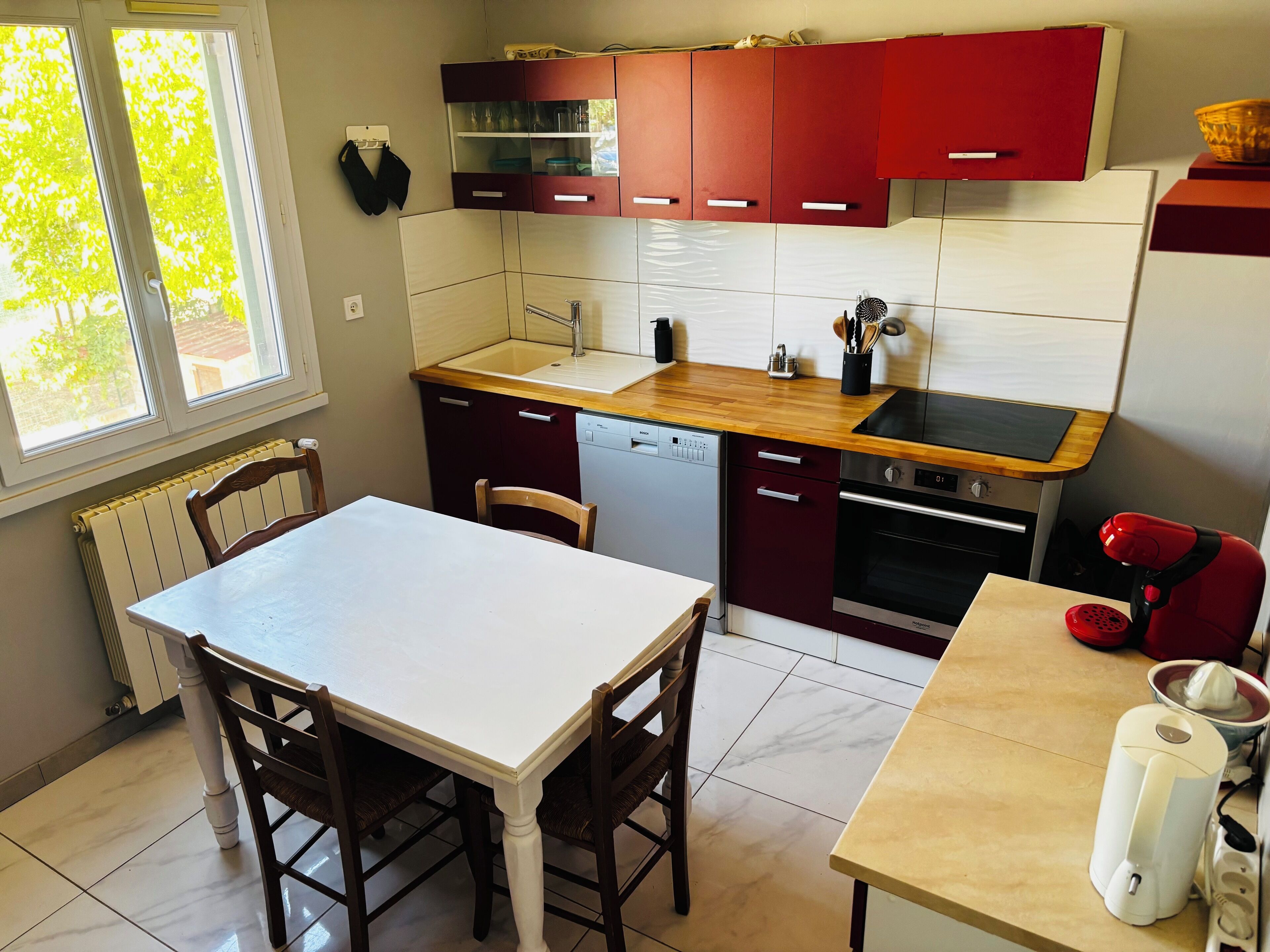 Een kleine keuken met een witte tafel, houten stoelen, een rode en witte kast, een vaatwasser en een rode stoel.