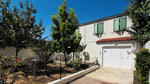 A white building with green shutters, a white garage door, and a paved driveway leading to a gated entrance. - 6-person house with garden in quiet neighborhood (Cournonterral)