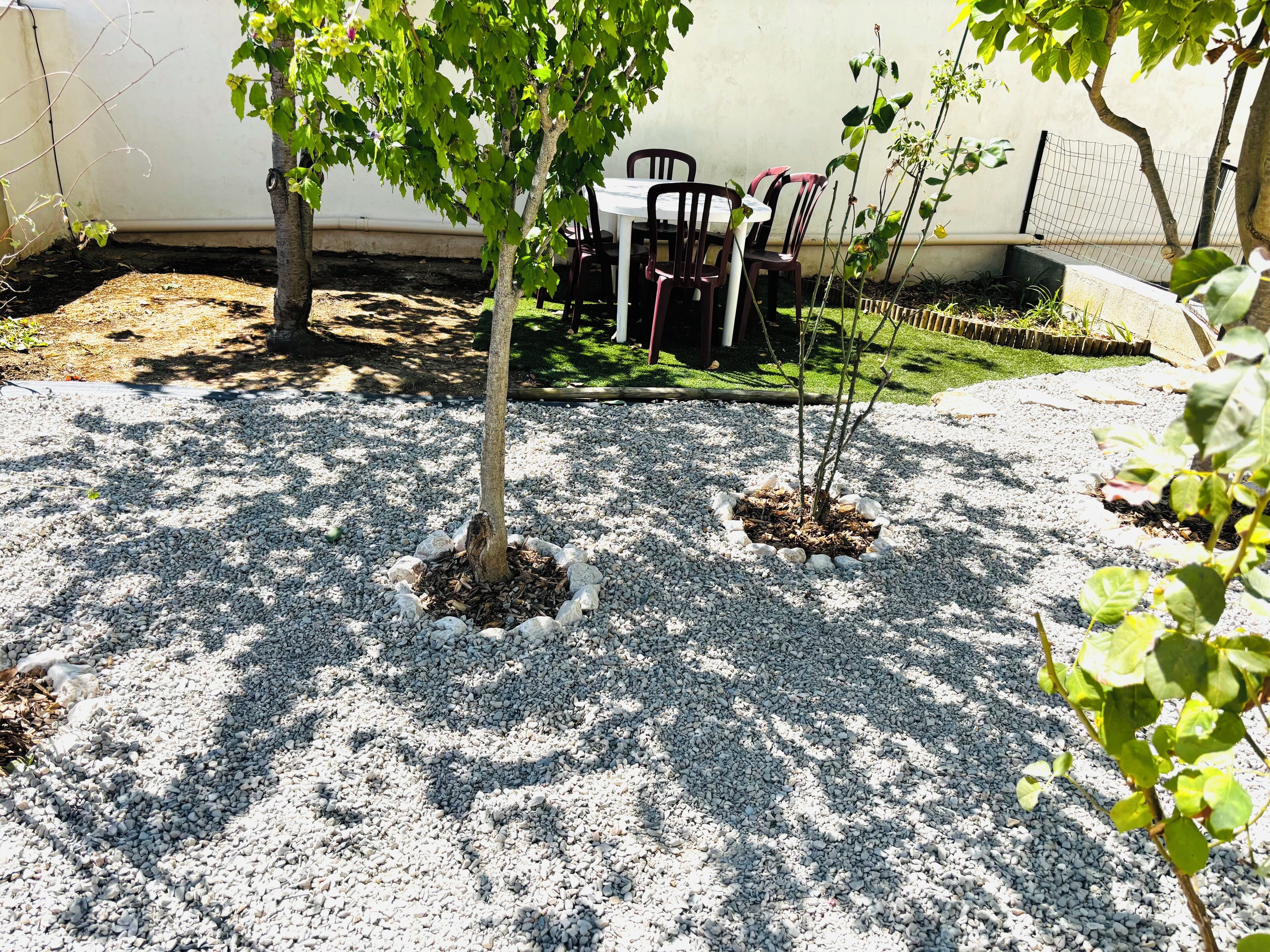 A patio area with a table and chairs, surrounded by gravel and small trees.