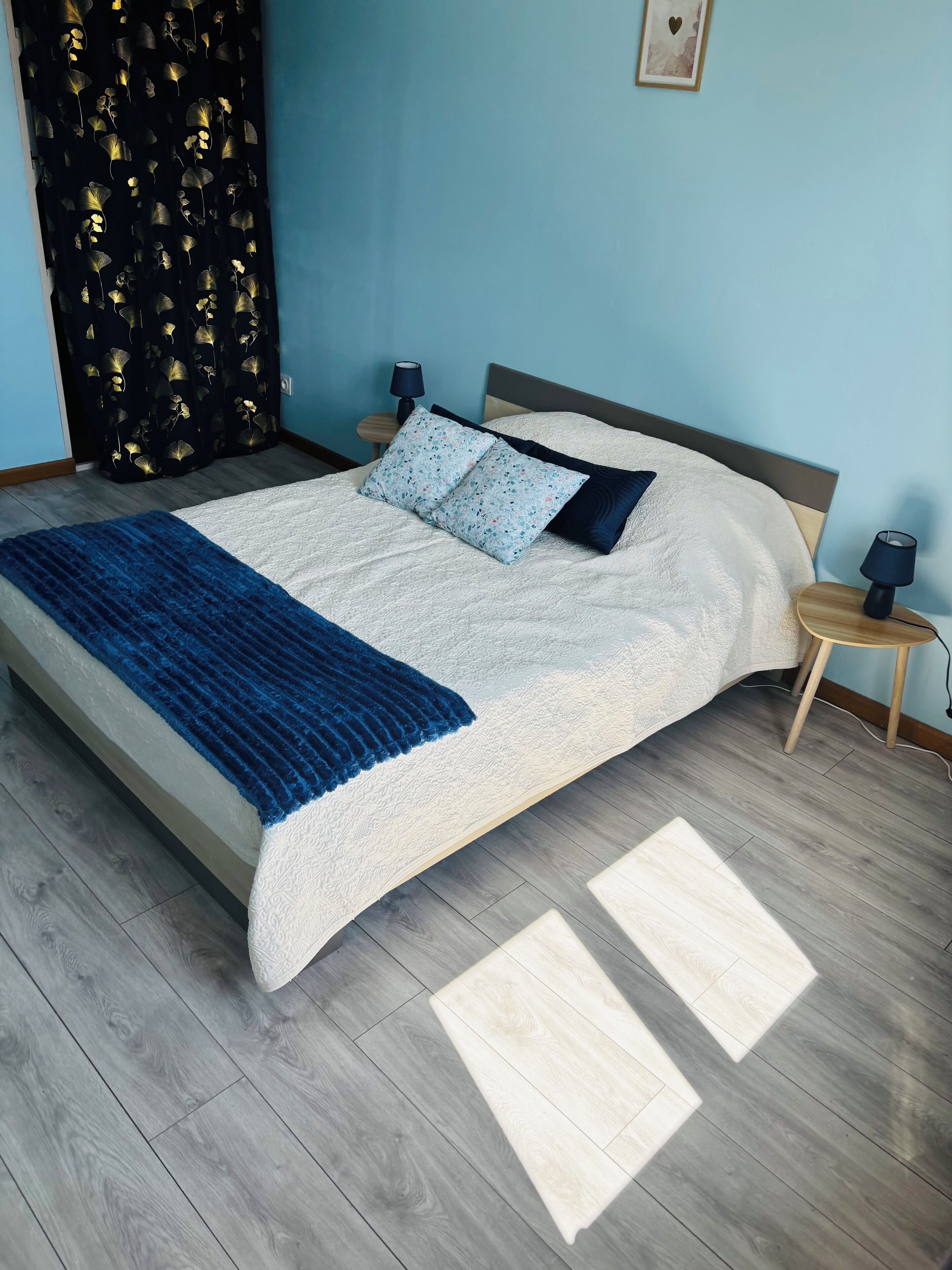 A neatly made bed with a blue blanket, two pillows, and a bedside table with a lamp.