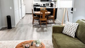 A modern living room with a green sofa, a brown ottoman, and a dining area with a round table and chairs. - Green Cottage / Explore Trails, Ride Bikes & Golf! (Bella Vista)