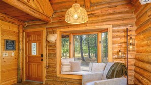 L’intérieur chaleureux d’un chalet en rondins, avec une porte en bois, un banc placé près d’une grande fenêtre donnant sur une forêt, et un luminaire suspendu.