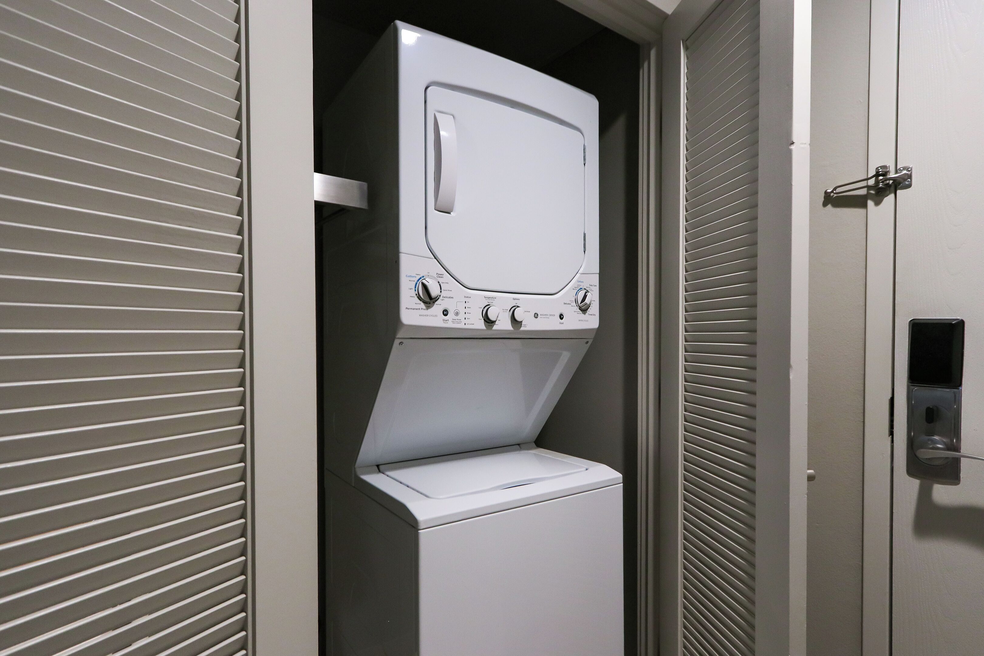 Een compact was- en droogapparaat, gemonteerd aan de muur, met bedieningspaneel, in een smalle ruimte met een deur en luiken.