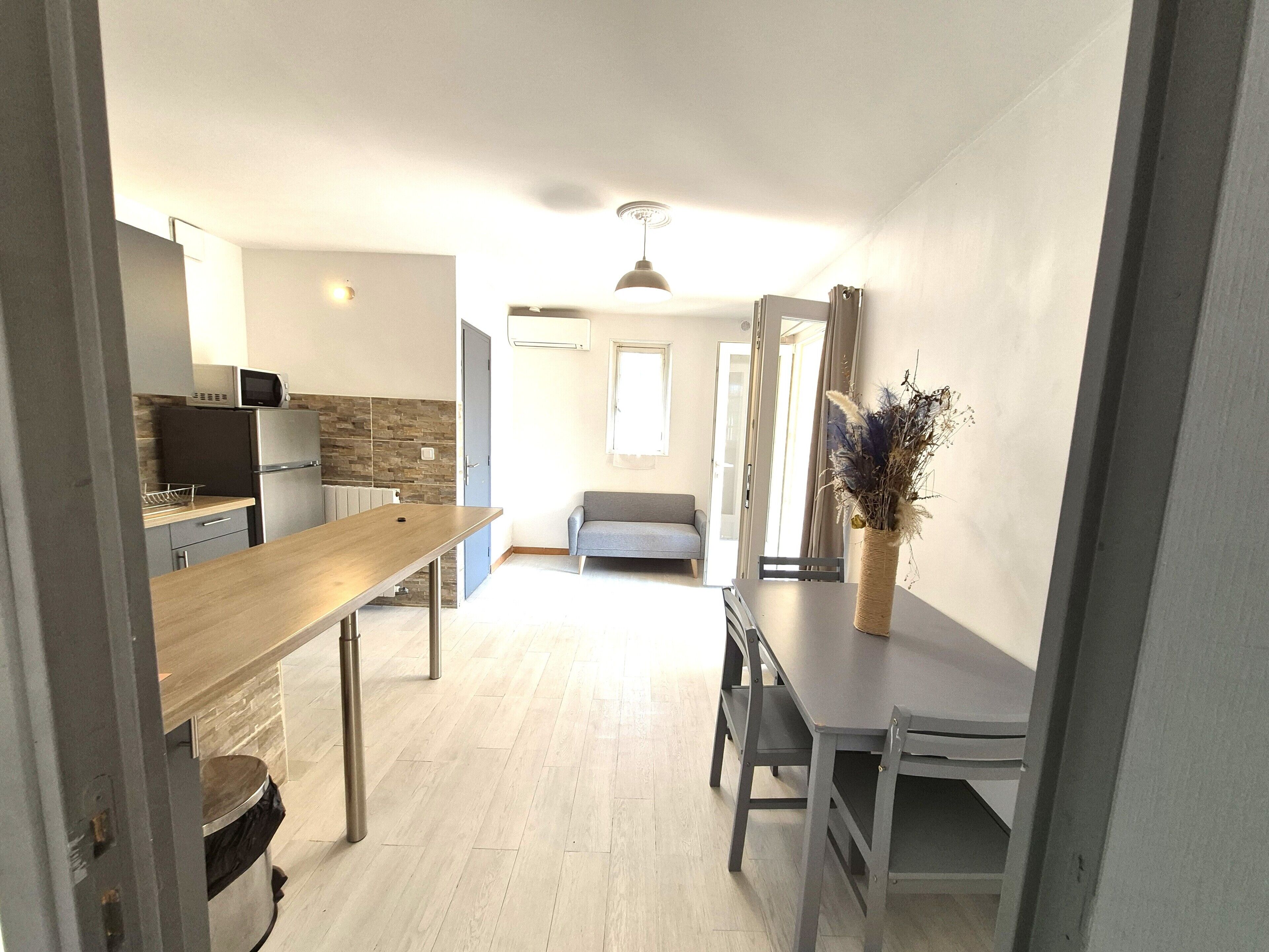 A modern kitchen with a wooden countertop, a built-in microwave, and a refrigerator. There is a dining table with chairs in the center of the room, and a gray sofa in the background.