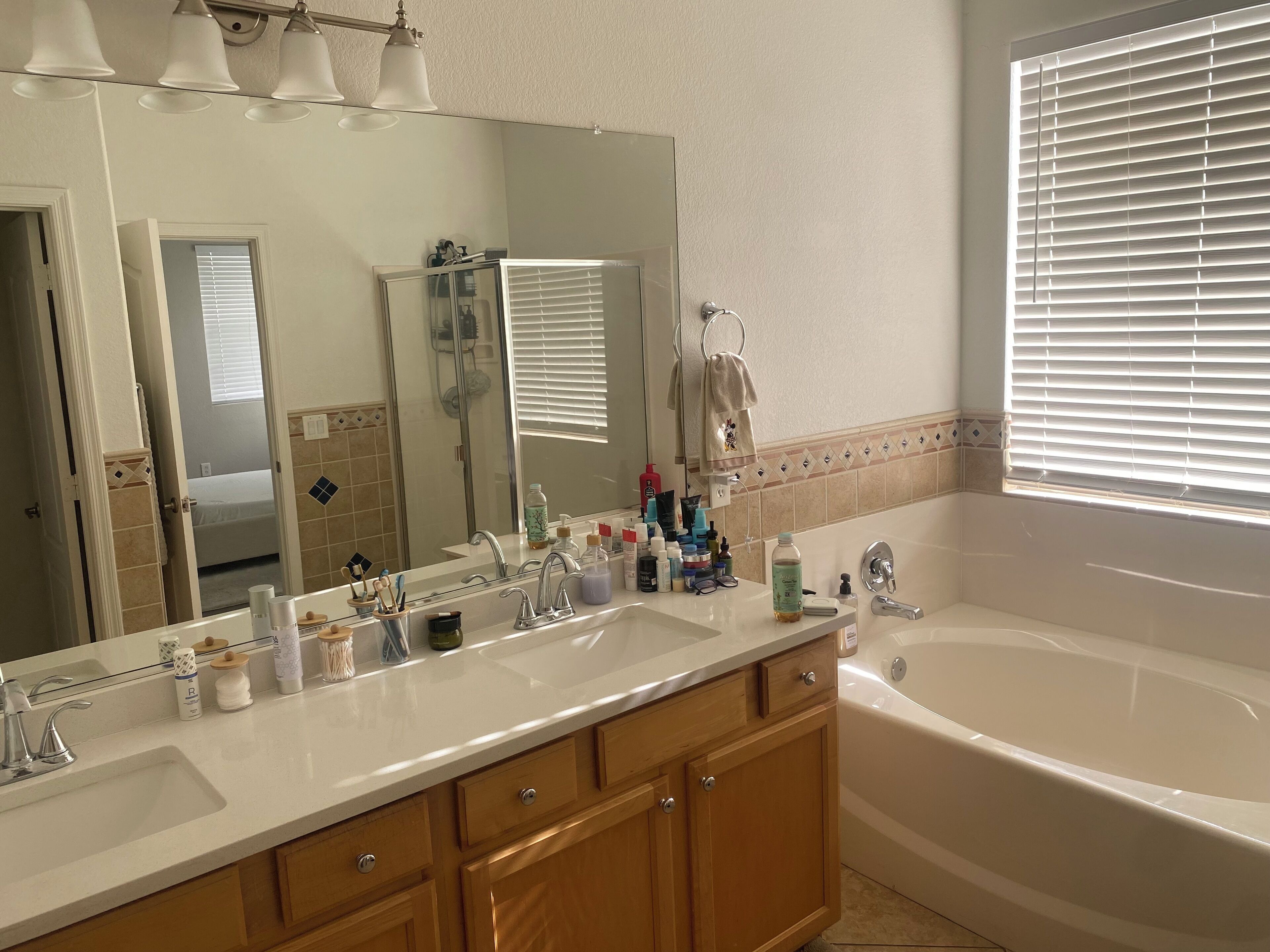 A bathroom with a large mirror, two sinks, a bathtub, and a window with blinds.