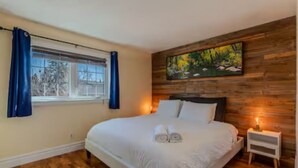 A bedroom with a large bed, wooden paneling on the walls, a painting above the bed, and a window with curtains. - Large Family-Friendly 3BE Home | Easy Access to QE2 (Airdrie)