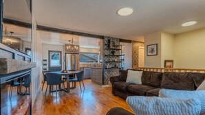 A spacious living area with a wooden floor, a dark brown sofa, and a dining table with chairs. - Large Family-Friendly 3BE Home | Easy Access to QE2 (Airdrie)