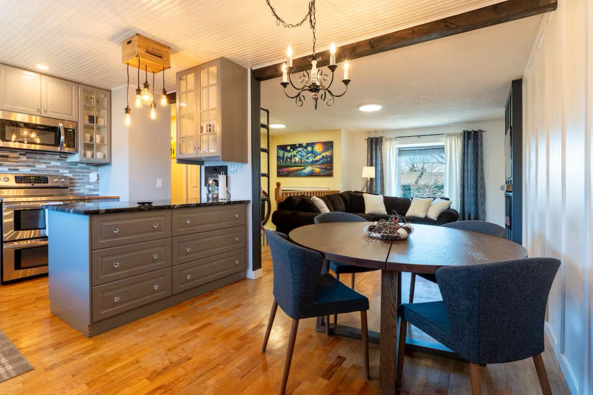 A modern kitchen with a central island, a dining area with a round table and chairs, and a living room with a sofa and a painting on the wall.
