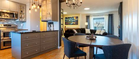 A modern kitchen with a central island, a dining area with a round table and chairs, and a living room with a sofa and a painting on the wall.