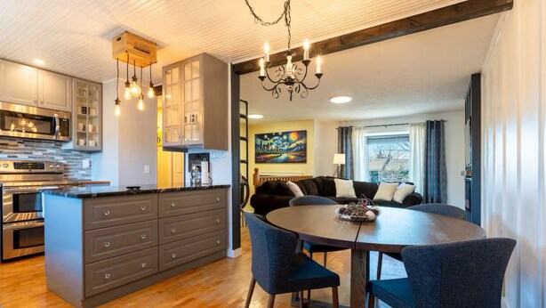 A modern kitchen with a central island, a dining area with a round table and chairs, and a living room with a sofa and a painting on the wall.