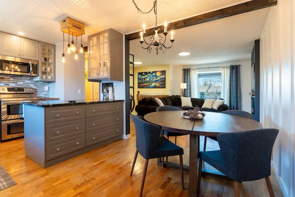 A modern kitchen with a central island, a dining area with a round table and chairs, and a living room with a sofa and a painting on the wall.