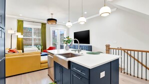 A modern kitchen with a central island, a dining area with a yellow sofa, and a staircase leading to an upper level. - Wharfside Whimsy | 3BR Retreat Walk to River St (Savannah)