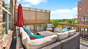 A balcony with wicker furniture, a red umbrella, and a view of greenery. - Wharfside Whimsy | 3BR Retreat Walk to River St (Savannah)
