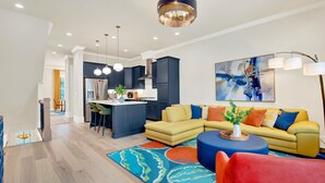 A modern living room with a yellow sectional sofa, a blue ottoman, and a colorful rug. There is a kitchen in the background with dark cabinets and a central island. - Wharfside Whimsy | 3BR Retreat Walk to River St (Savannah)