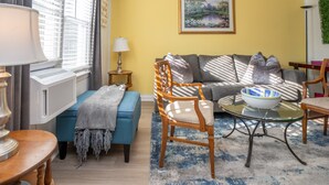 Una sala de estar con un sofá azul, una silla de madera, una mesa de centro de vidrio y un cuadro en la pared.
