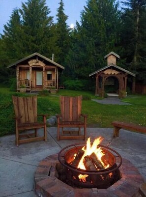 A wooden cabin with a fire pit in the foreground, surrounded by a grassy area and trees. - Bond Ranch Retreat, Rustic Olympic Peninsula (Sequim)