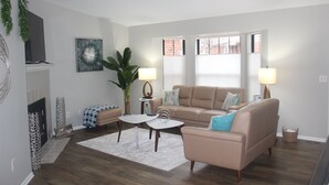 Una sala de estar con un sofá color beige, dos mesitas de noche con lámparas, una mesa de centro, una chimenea y un ventanal grande con persianas.