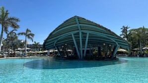 Una estructura amplia y abierta, con un diseño de techo singular, rodeada por una piscina cristalina y palmeras.