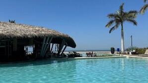 Piscina azul y cristalina con una estructura de techo de paja, palmeras y vista a la playa.