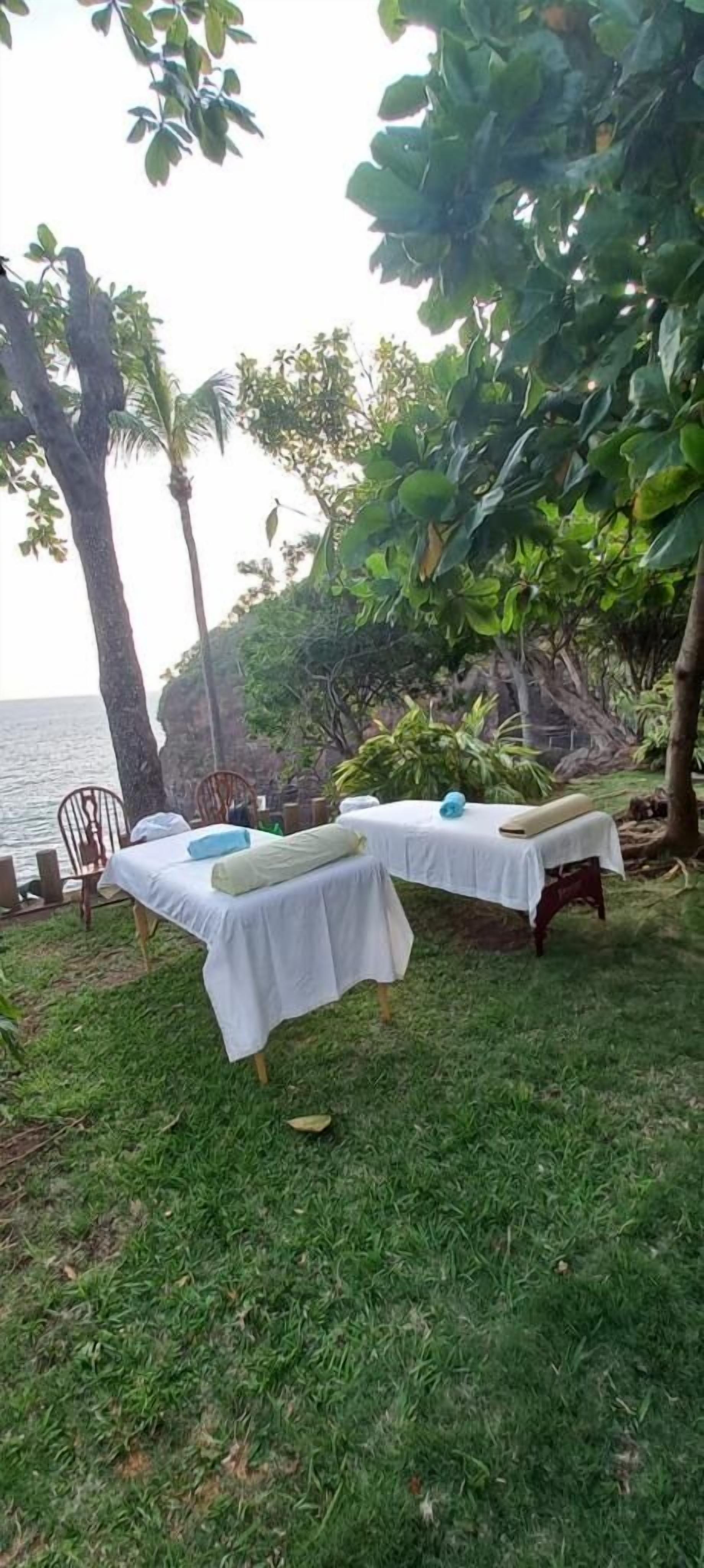 Two outdoor tables set up on a grassy area with chairs, surrounded by trees and a view of the sea.