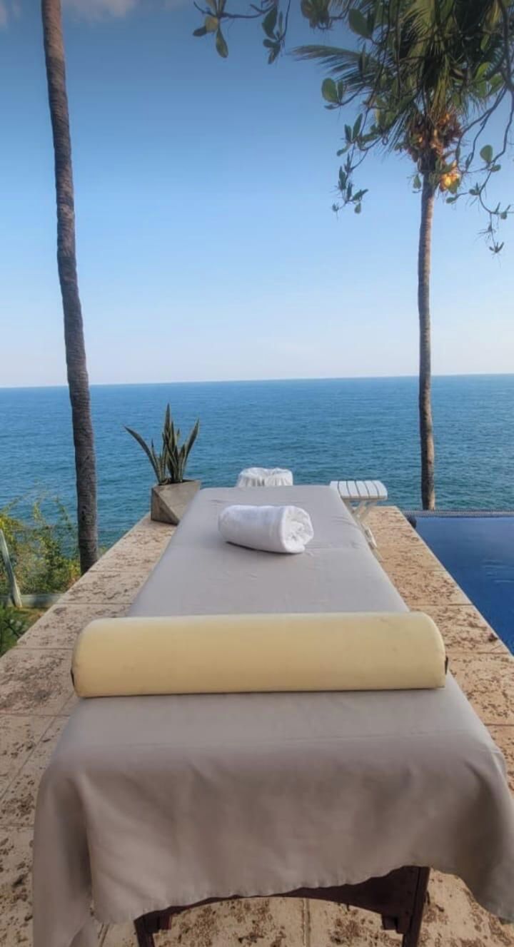 A massage table with a towel, set against a backdrop of a clear sky and ocean view, with palm trees and a potted plant nearby.