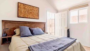 Un dormitorio con cabecera de madera, una gran obra de arte tejida en la pared, una cama con colcha color beige y dos almohadas, una mesita de noche con lámpara, y una ventana que da a un muro de ladrillo.