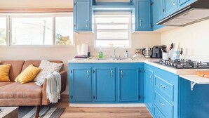 Una cocina con armarios azules, una encimera blanca y una estufa. Hay una ventana sobre el fregadero y un sofá marrón en la sala de estar.