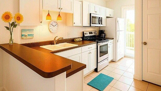 A kitchen with white cabinets, a microwave, stove, sink, and a clock. There are two hanging lights and a vase with flowers on the counter.
