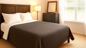 A neatly made bed with a dark quilt, a wooden headboard, a bedside table with a lamp, and a dresser with a mirror.
