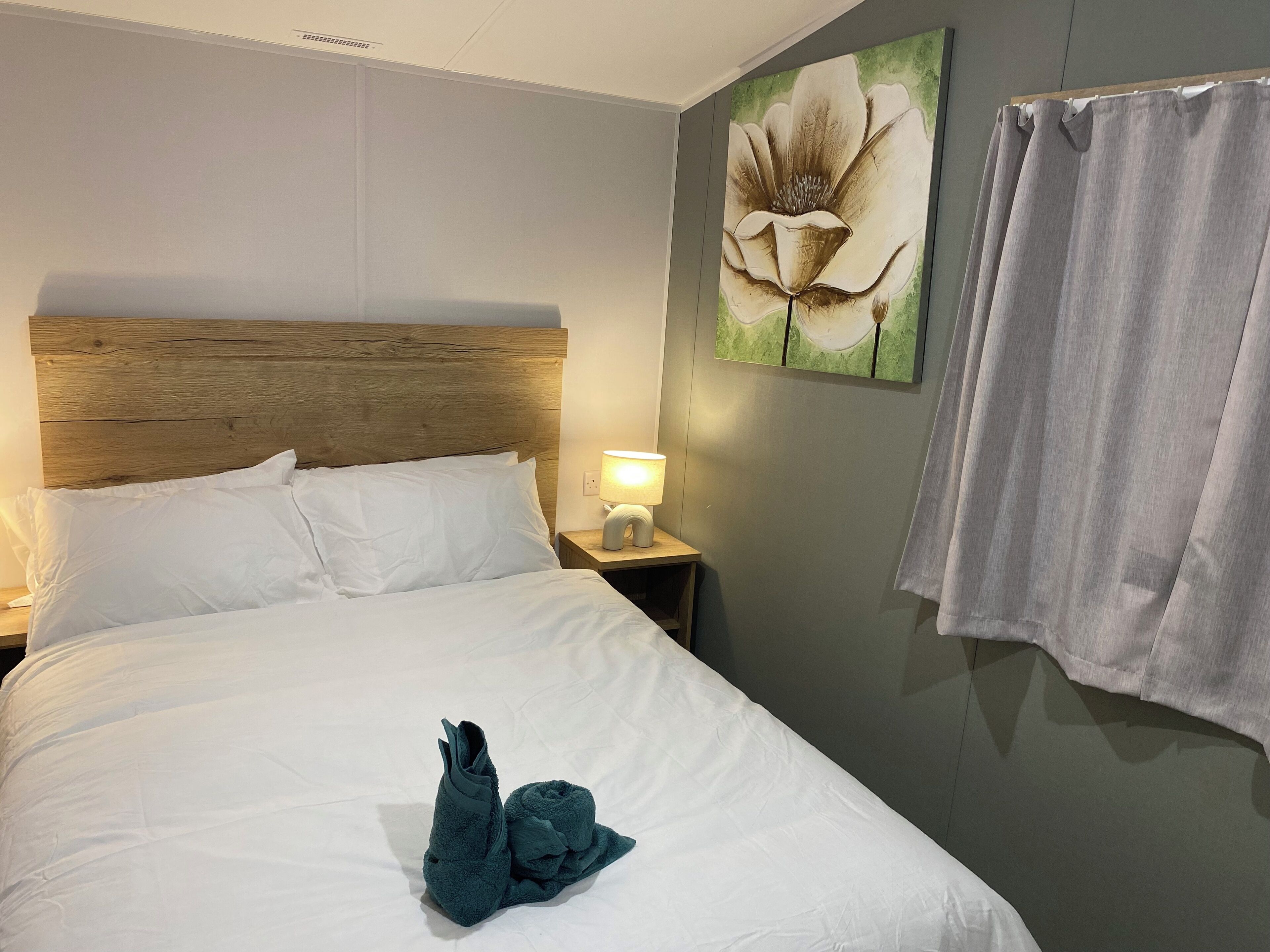 Ein ordentlich bezogenes Bett mit weißen Leinen, ein Holz-Kopfbrett, eine Nachttischlampe und ein Blumenbild an der Wand.