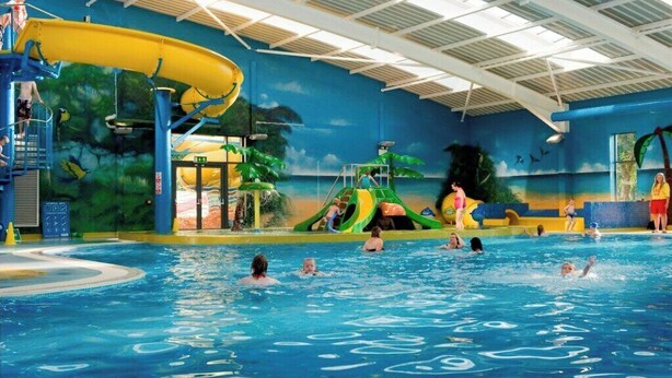 En inomhuspool med en gul vattenrutschkana, en lekplats för barn och en väggmålning som föreställer en tropisk miljö.