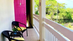 A balcony with two wicker chairs, a pink door, and a view of greenery. - Villa Mia Apartments Studio #3 (Oistins)
