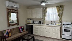 A compact living space with a kitchenette, a sofa with colorful cushions, a glass coffee table, and a ceiling fan. - Villa Mia Apartments Studio #3 (Oistins)