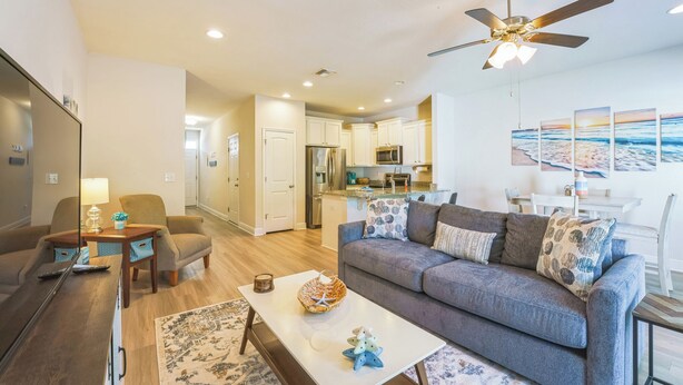 A modern living room with a grey sofa, a coffee table, and a TV. There is a kitchen in the background with white cabinets and a microwave. - Bright & Modern Beach Getaway-Pool+Prime Location (Panama City Beac)