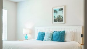 A neatly made bed with white and blue pillows, a framed picture of palm trees on the wall, and a bedside lamp. - Bright & Modern Beach Getaway-Pool+Prime Location (Panama City Beac)