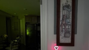Un pasillo con un cuadro enmarcado de un oso sobre un banco, una luz con forma de corazón, y una vista parcial de un comedor con sillas y una mesa.