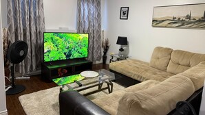 Una sala de estar con un televisor de pantalla plana, un sofá color beige, una mesa de centro de vidrio, un otoman de cuero negro y una alfombra blanca.
