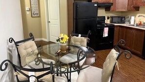 Un comedor con una mesa de vidrio y cuatro sillas, un refrigerador negro, un microondas y armarios de madera al fondo.