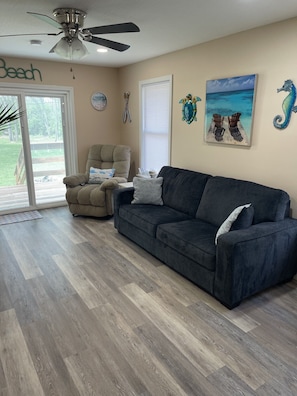 A living room with a dark grey sofa, a beige armchair, a ceiling fan, and a large window with a view of the outdoors. - Cozy 1-bedroom home near Circleville, Walking distance to Hargus Lake. (Circleville)