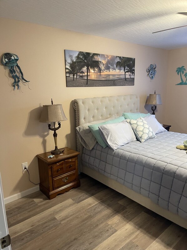 Une chambre à coucher avec un lit, des tables de chevet, une lampe fixée au mur et un tableau sur le thème de la plage.