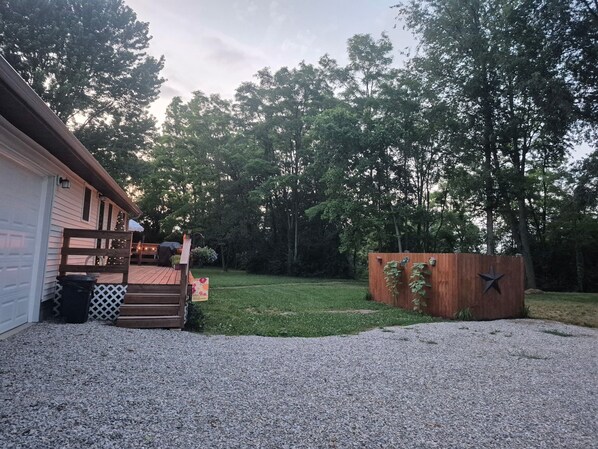 A residential property with a gravel driveway, a wooden deck, a garage, and a fenced yard with trees.