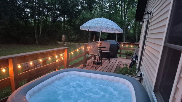 Een hottub op een houten terras met lichtjes, een tafel en stoelen, en een overdekte barbecueplaats.