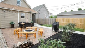 A backyard with a patio area featuring wooden tables and benches, a fire pit, and a barbecue grill. - Themed Home | Walk to Downtown | Arcade | Fire Pit (Indianapolis)