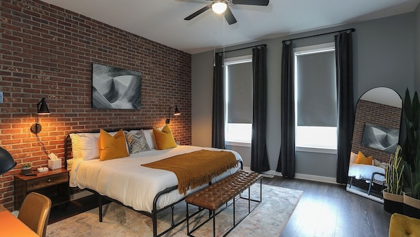 A modern bedroom with a brick wall, a bed with a bench, a ceiling fan, and a large window with curtains. - Themed Home | Walk to Downtown | Arcade | Fire Pit (Indianapolis)