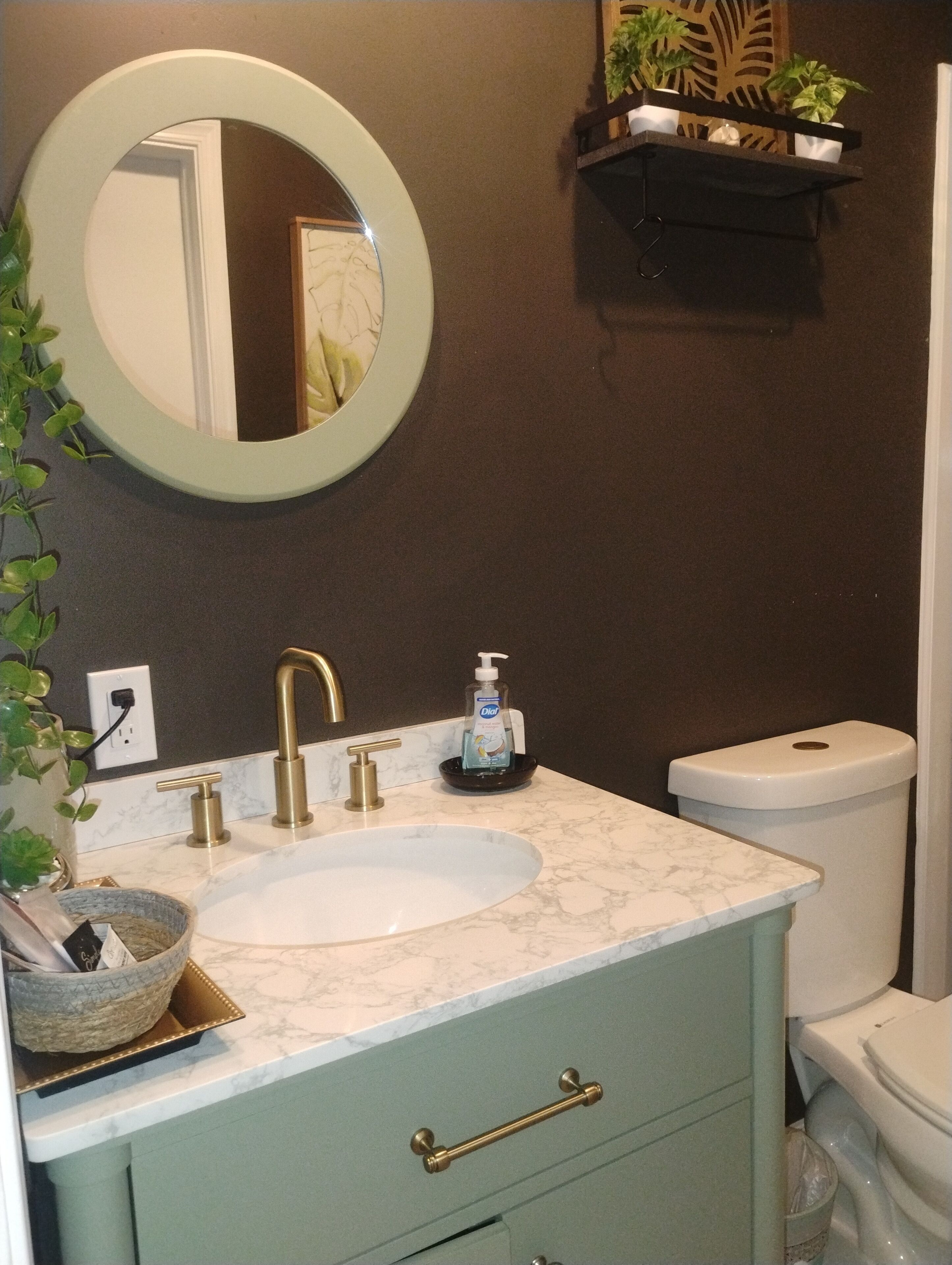 A bathroom with a white sink, gold faucets, a marble countertop, a round mirror, a toilet, and a small shelf with plants.