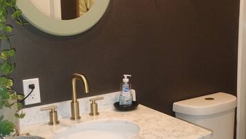 A bathroom with a white sink, gold faucets, a marble countertop, a round mirror, a toilet, and a small shelf with plants.