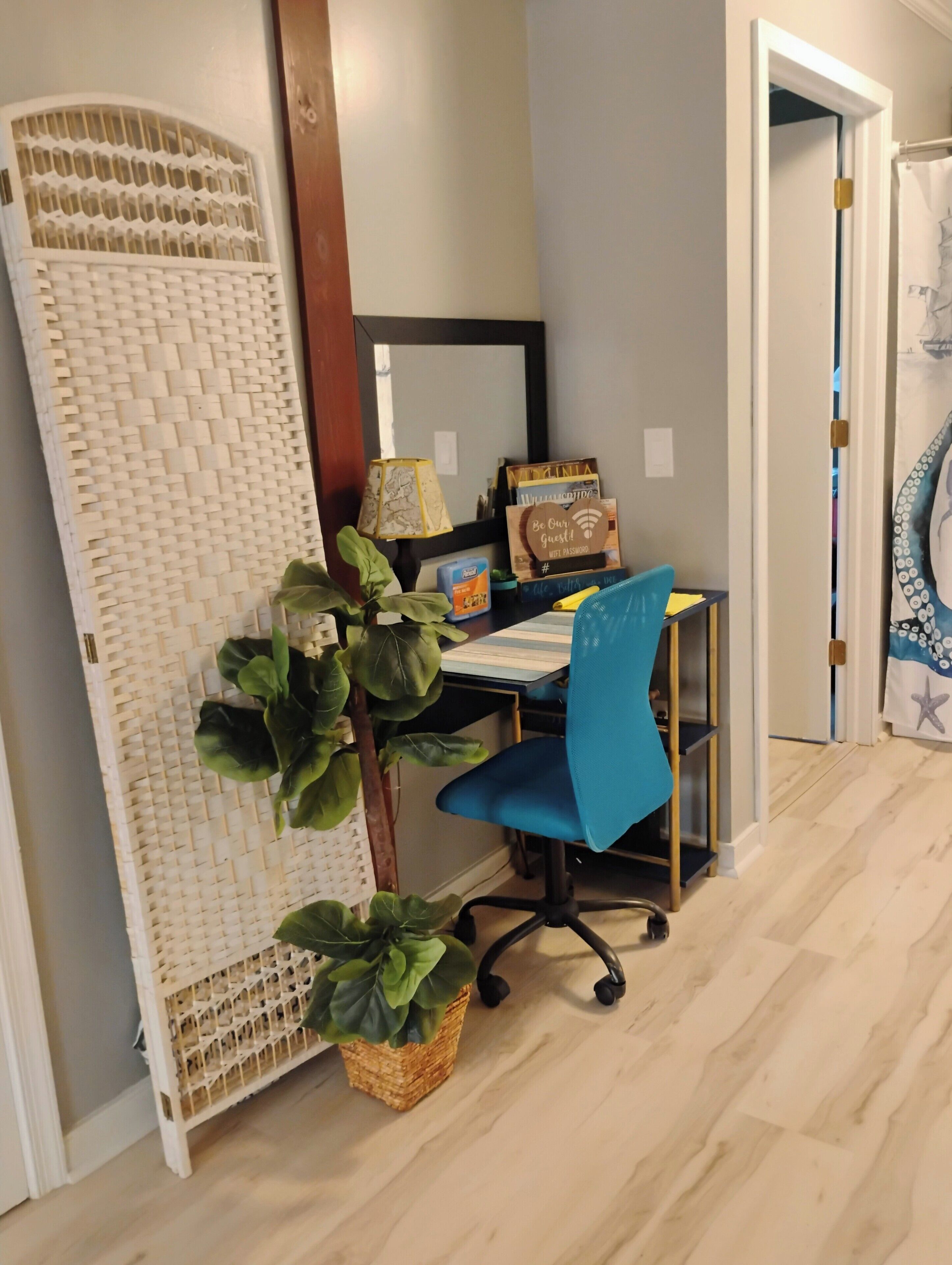 A room with a desk, chair, and a large woven partition. There are plants, a lamp, and a door with a curtain.
