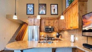 A kitchen with wooden cabinets, a granite countertop, a stainless steel refrigerator, a microwave, and a bar area with stools. - Mountain bikers dream near Gondola in Northstar (Truckee)